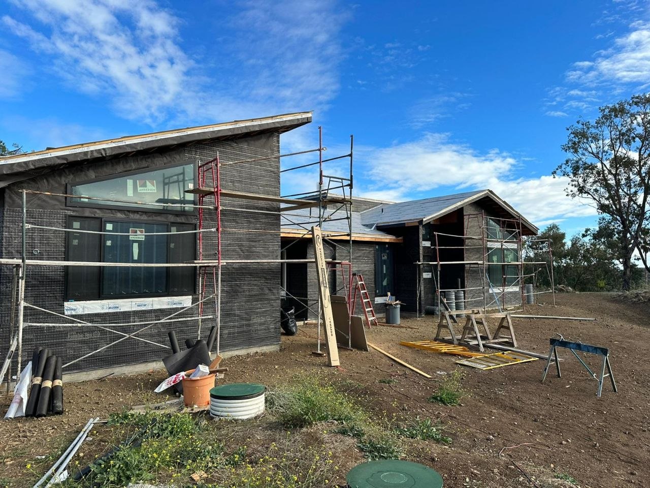 Modern L-shaped custom residential build mid-construction — shed cathedral roofline, dark stone veneer, Zip-System sheathing, Andersen windows, timber V-truss posts — PBG, Los Angeles.