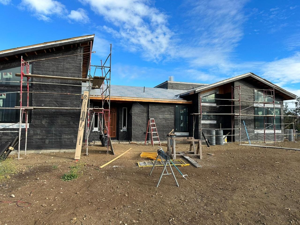 Front elevation of a modern L-shaped custom residential build mid-construction — dark stone veneer, cathedral roofline, scaffolding wrap — PBG, Los Angeles.