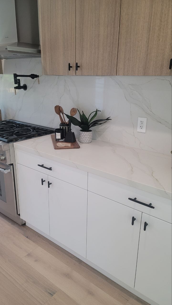 Modern white kitchen — pro gas range with pot filler, Calacatta-vein quartz backsplash, light-oak upper cabinets and matte-white lower cabinets over wide-plank white-oak floors — PBG custom residential build, Los Angeles.
