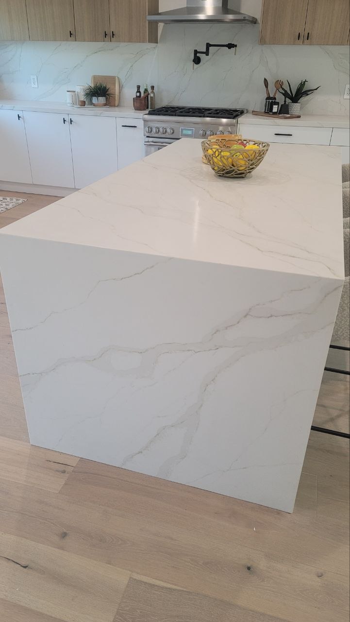 Waterfall-edge Calacatta-vein quartz island with matte-black hardware, brass bowl accent, and light-oak upper cabinets — PBG custom kitchen, Los Angeles.