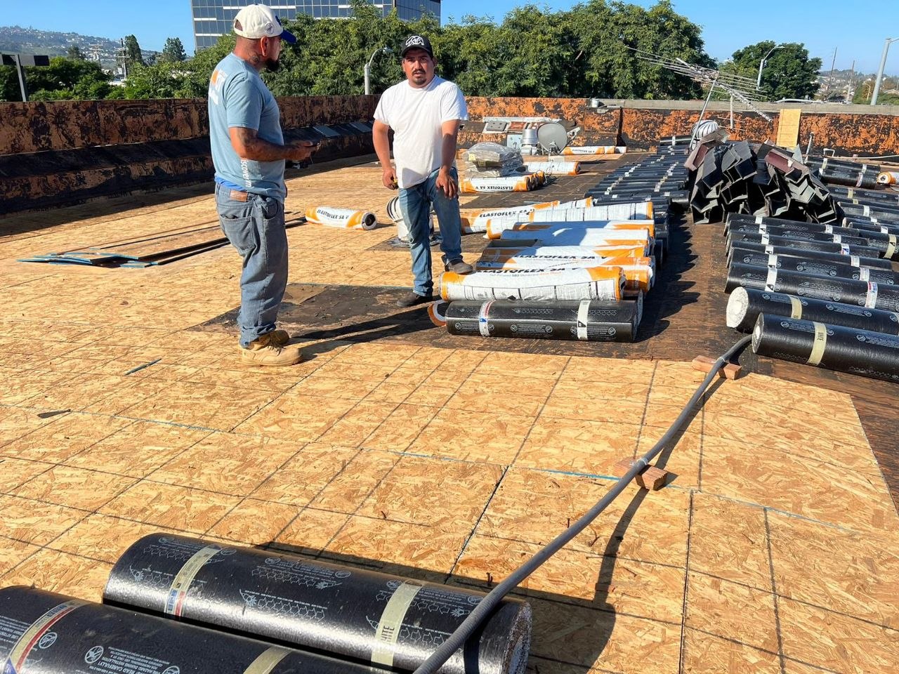 PBG crew staging Elastoflex SA V modified-bitumen membrane rolls on a new OSB commercial flat-roof deck with hillside Los Angeles context — process shot of a PBG commercial roofing project.