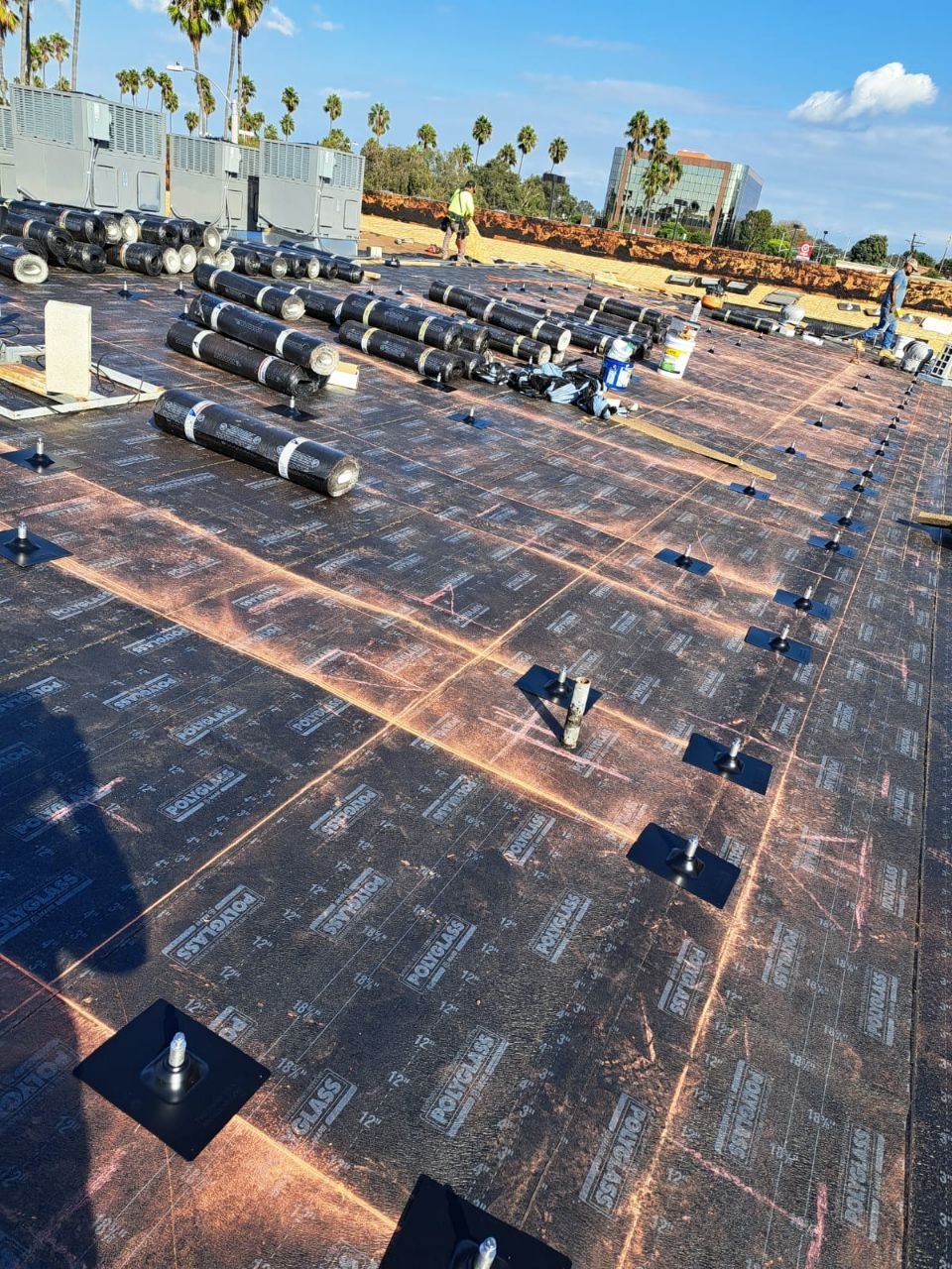 Wide overview of a Polyglass modified-bitumen commercial flat-roof install — base sheet in place with fasteners set, cap sheet rolls staged, Los Angeles skyline in the background — PBG commercial roofing.
