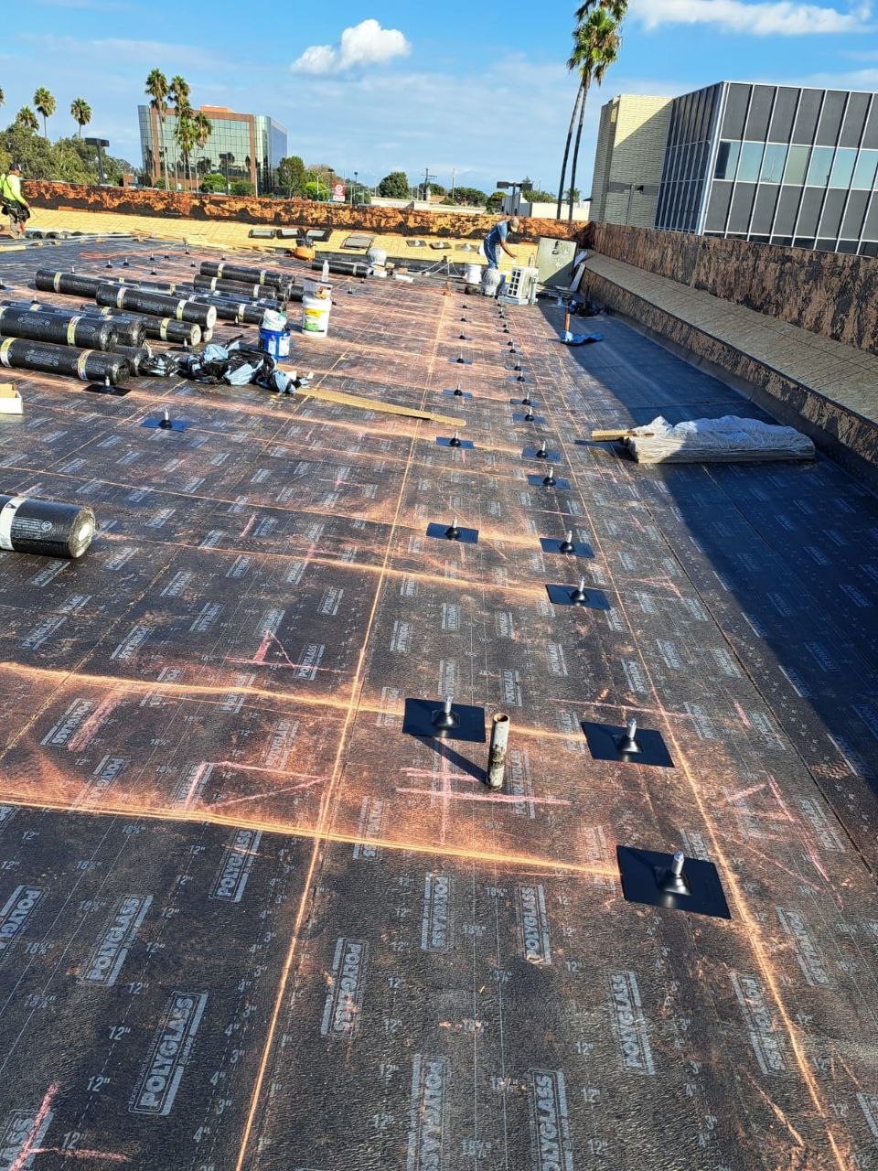 Polyglass modified-bitumen base sheet installed with fastener pattern, cap sheet rolls staged toward the parapet with neighboring building context — PBG commercial roofing.