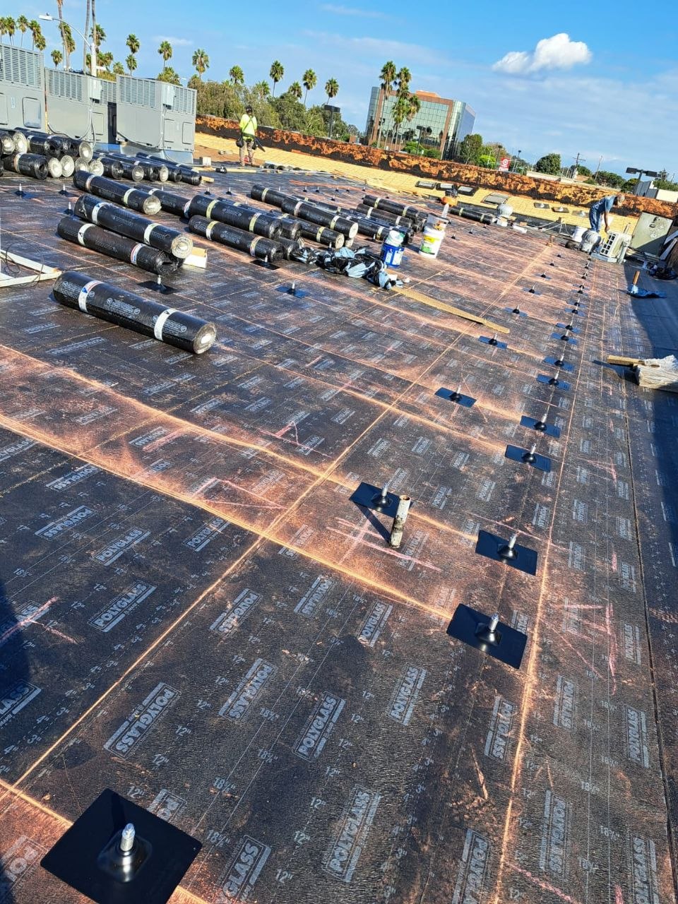Polyglass modified-bitumen cap sheet rolls staged in a precision grid pattern over the installed base sheet with visible fastener plates — PBG commercial roofing craft detail.