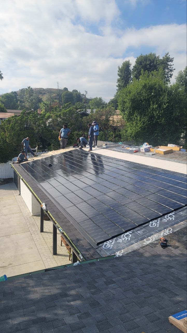 Wide view of a completed BIPV solar-integrated roof next to conventional composition shingle for contrast, with the install crew finishing edges — PBG custom residential build, Los Angeles.