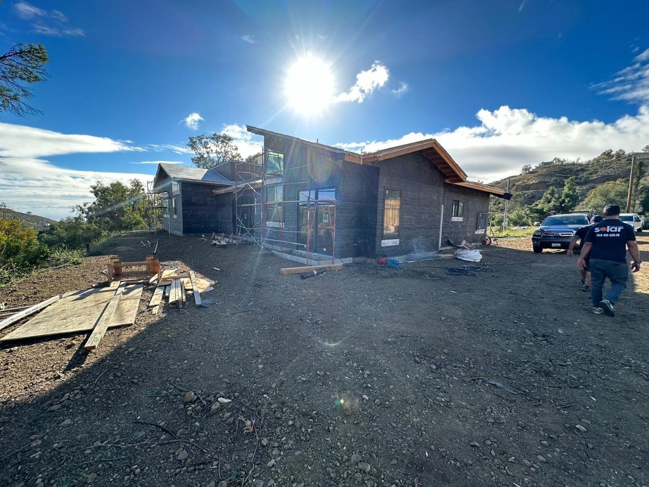 Modern custom residential build mid-construction with solar crew arriving — hilltop backdrop, wood siding, cathedral roofline — PBG, Los Angeles.